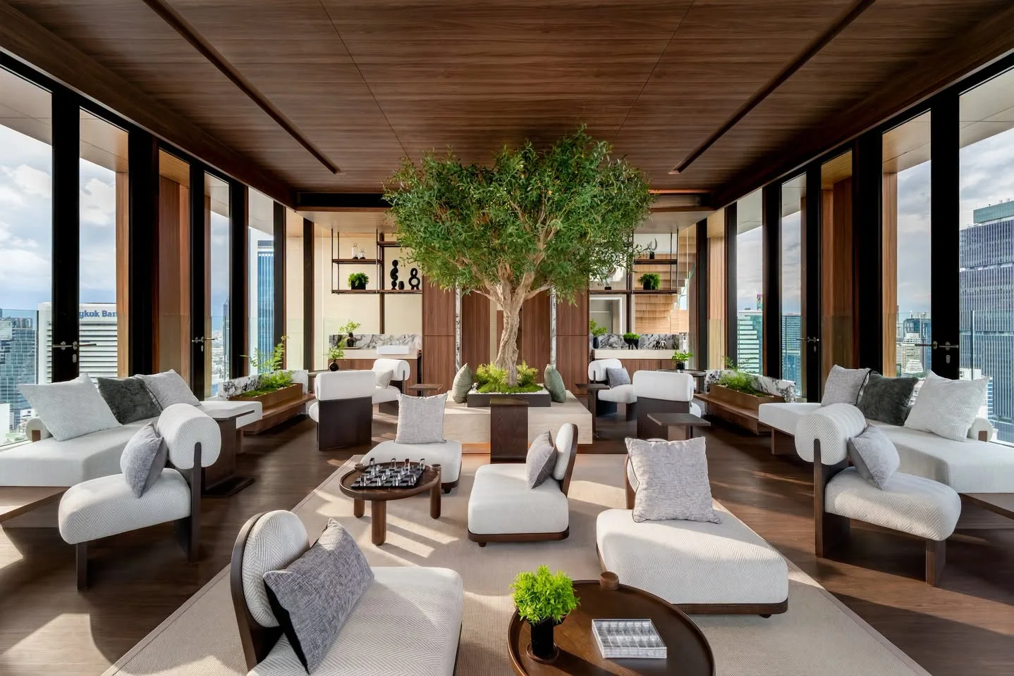 A high-floor lounge with an olive tree, white armchairs and windows over the city